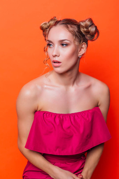 The girl in a red dress on a orange background in the studio. Blonde girl with two hair knots holding hands on knees, looking left - Foto, afbeelding