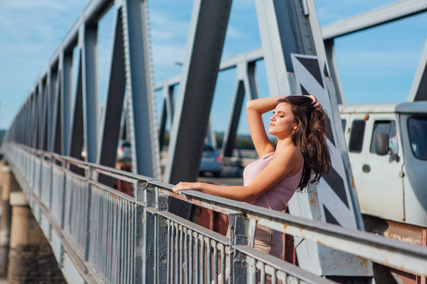 Warm summer portrait of a young beautiful woman posing on the old rusty transport bridge over the river during sunset. - Фото, изображение