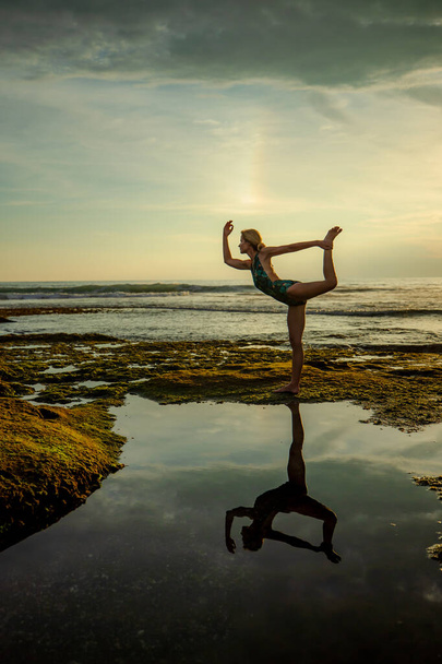 Outdoor sunset yoga. Attractive woman practicing yoga, standing in Natarajasana, Lord of the Dance Pose. Amazing water reflection. Bali, Indonesia - 写真・画像