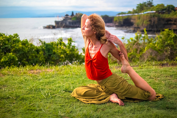 Trening jogi na świeżym powietrzu. Młoda kobieta ćwicząca Eka Pada Rajakapotasana, Jednonogi King Pigeon Pose. otwieracz poprawia oddychanie. Świątynia Tanah Lot, Bali, Indonezja - Zdjęcie, obraz