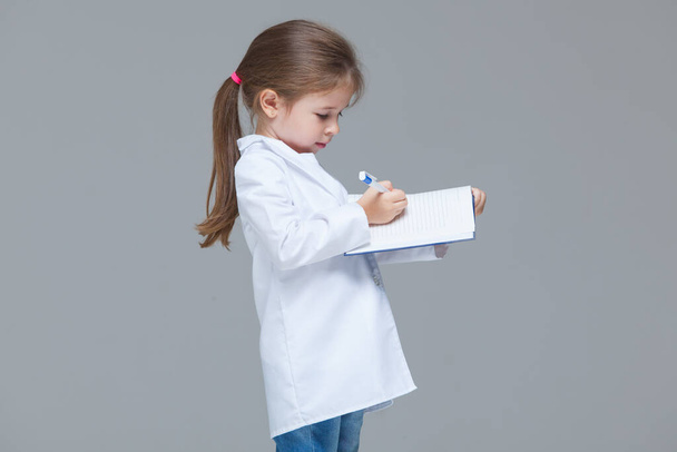 Adorable child girl uniformed as doctor is writing medical record isolated on grey background - Foto, Imagem