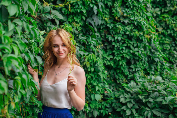 Portrait of a young beautiful woman with make up and curly blond hair, standing next to the wall of wild grape leaves. Copy space. - Foto, Imagen
