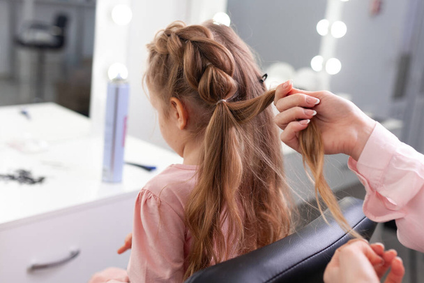 The work of the stylist on hairstyles in a bright salon, the girl makes curls and braids braids of a small beautiful model, indoor - Foto, immagini