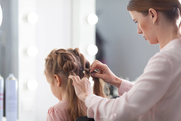 The work of the stylist on hairstyles in a bright salon, the girl makes curls and braids braids of a small beautiful model, indoor - Foto, afbeelding