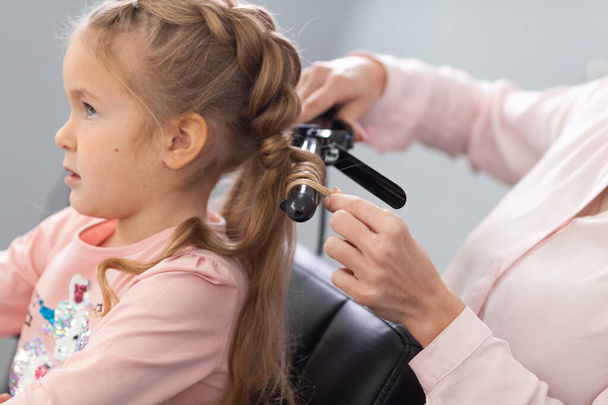 The work of the stylist on hairstyles in a bright salon, the girl makes curls and braids braids of a small beautiful model, indoor - Photo, Image