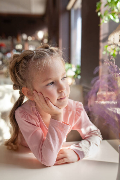 Belle petite fille en robe rose assise sur un canapé blanc dans un café, journée ensoleillée et bel intérieur violet vintage, fille regardant par la fenêtre
 - Photo, image