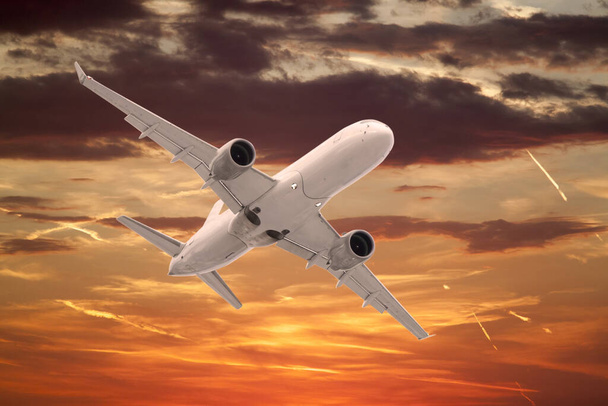 white passenger plane flies against on background of beautiful sunset sky is orange with clouds and condensation traces - Photo, Image