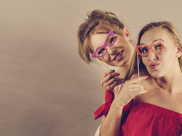 Happy two women holding paper decoration photo booth mask glasses on stick, having fun. Wedding, birthday and carnival funny accessories concept. - Φωτογραφία, εικόνα
