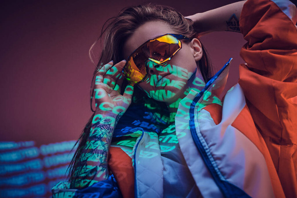 Tattooed racer girl standing over neon vivid text projection in a bright studio holding her glasses, close up look - 写真・画像