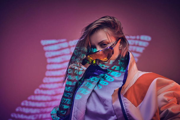 Tattooed racer girl standing over neon vivid text projection in a bright studio holding her glasses, close up look - Photo, Image