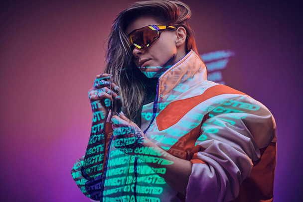 Tätowiertes Rennfahrermädchen steht über neonfarbener Textprojektion in einem hellen Studio und hält ihre Brille, Großaufnahme - Foto, Bild