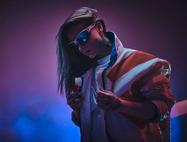 Neon renkli canlı bir stüdyoda duran dövmeli yarışçı kız, güneş gözlüğü takarak havalı ve riskli görünüyor. - Fotoğraf, Görsel
