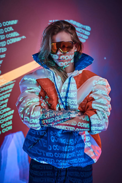 Татуированная гонщица стоит над неоновой яркой текстовой проекцией в яркой студии, держа очки, крупным планом
 - Фото, изображение