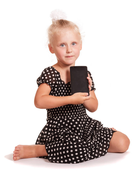 Little girl in a dark dress with polka dots, holding a mobile while sitting isolated on white background. - Foto, afbeelding