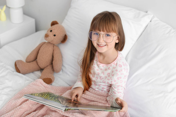 Aranyos kislány szemüveget visel, miközben könyvet olvas az ágyban - Fotó, kép