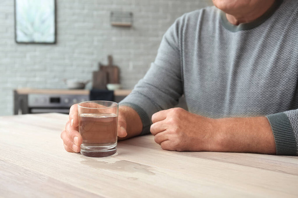 Vanhempi mies, joka kärsii Parkinsonin oireyhtymästä lasillisella vettä kotona
 - Valokuva, kuva