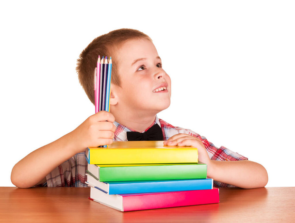Szczęśliwy uczeń z ołówkami i książkami siedzi przy stole odizolowany na białym tle. - Zdjęcie, obraz