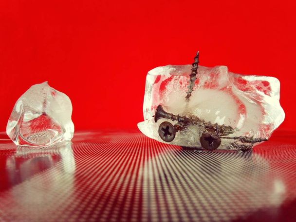 A cold shard of ice with metal screws frozen in it on a textured surface and a red background in a creative macro photo - Zdjęcie, obraz