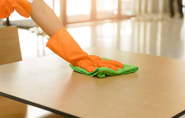 Γυναικεία χέρια σε προστατευτικά γάντια πορτοκαλί καουτσούκ που κρατούν πράσινο πανί καθαρισμού μικροϊνών και σκουπίστε τη σκόνη χρησιμοποιώντας ένα διάλυμα αποστείρωσης ψεκασμού κάνουν τον καθαρισμό και την απολύμανση για καλή υγιεινή - Φωτογραφία, εικόνα