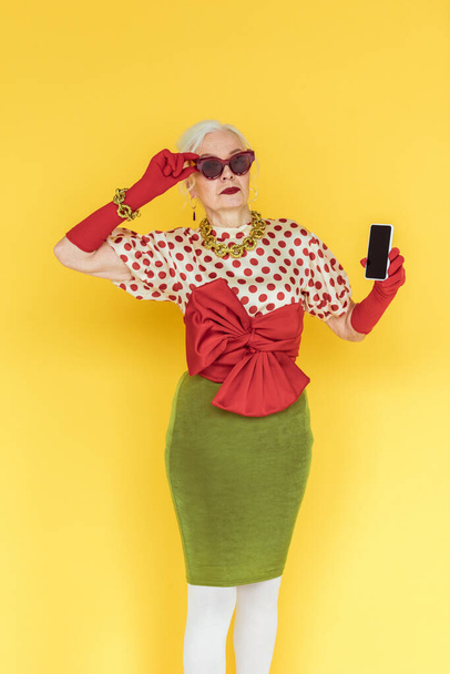 Stylish senior woman in sunglasses holding smartphone on yellow background - Photo, Image