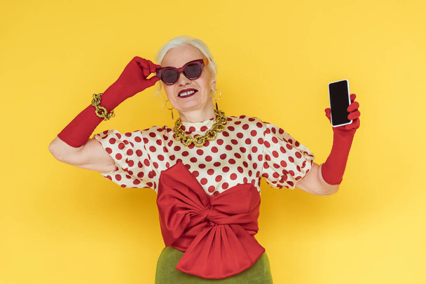 Stilvolle Seniorin lächelt und hält Smartphone mit leerem Bildschirm auf gelbem Hintergrund - Foto, Bild