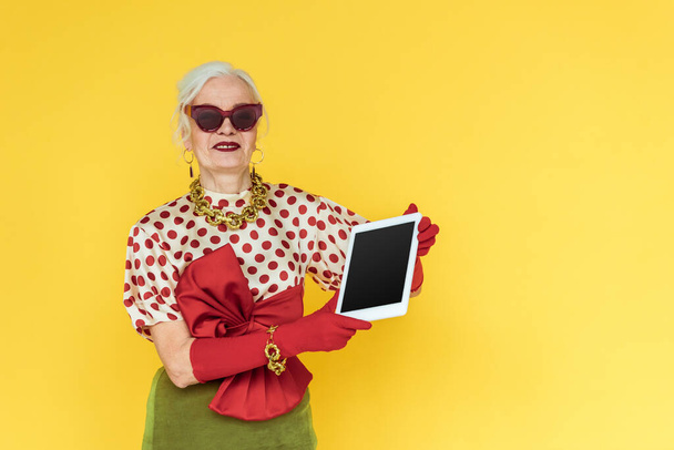 Mujer mayor de moda sonriendo mientras sostiene la tableta digital aislada en amarillo
 - Foto, Imagen