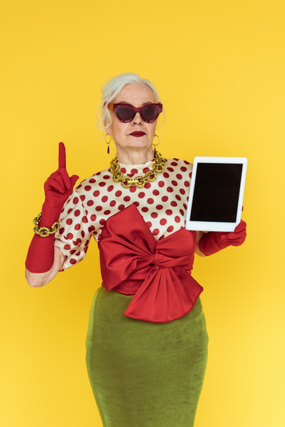 Modieuze oudere vrouw in een zonnebril met digitale tablet en wijzend met de vinger geïsoleerd op geel - Foto, afbeelding