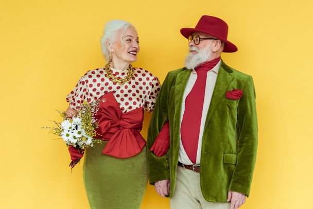 Stylish senior couple with bouquet of wildflowers smiling at each other on yellow background - Foto, immagini