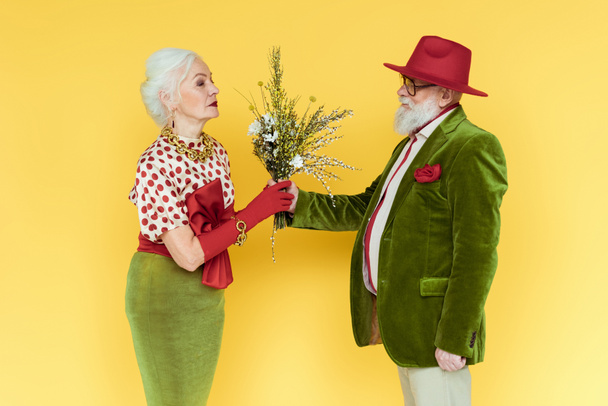 Side view of stylish senior man giving wildflowers to beautiful woman on yellow background - 写真・画像