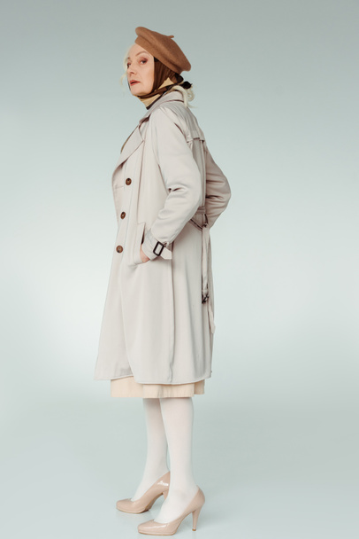 Sivunäkymä tyylikäs vanhempi nainen trenssi takki katsomalla kameraa valkoisella taustalla
 - Valokuva, kuva