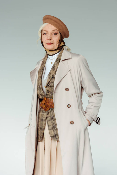 Fashionable senior woman in beret and trench coat looking at camera isolated on grey - Foto, imagen