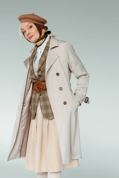Elegant senior woman with hand in pocket of trench coat looking at camera isolated on grey - Foto, immagini