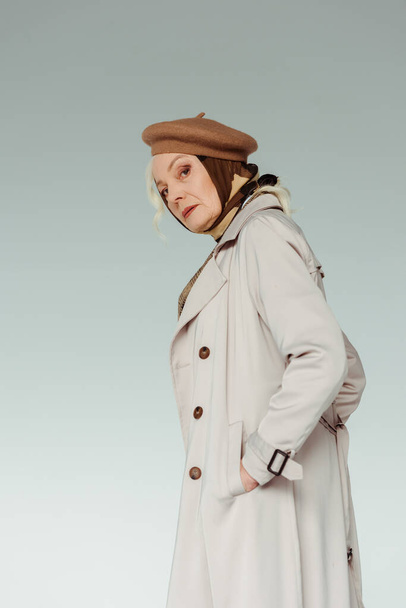 Side view of stylish senior woman looking at camera while holding hand in pocket of coat isolated on grey - Foto, Imagem