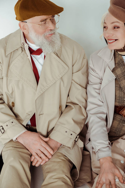Stylish senior couple smiling at each other while sitting on white surface on grey background - Foto, afbeelding