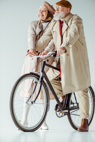 Volle Länge einer eleganten Seniorin, die in der Nähe eines gutaussehenden Mannes auf einem Fahrrad auf weißem Hintergrund weglächelt - Foto, Bild