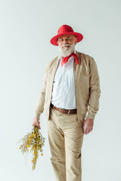 Fashionable elderly man in red hat holding wildflowers and smiling at camera isolated on white - Photo, image