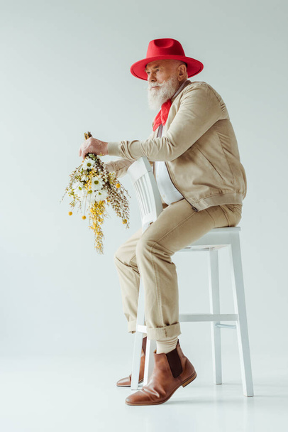 Side view of handsome senior man in red hat holding wildflowers while sitting on chair on white background - Photo, Image