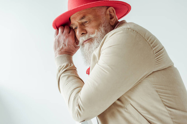 Vista lateral de un hombre mayor con estilo en sombrero rojo mirando hacia otro lado mientras está sentado en la silla aislado en blanco
 - Foto, imagen