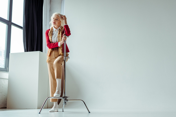 Όμορφη ηλικιωμένη γυναίκα κοιτάζει μακριά κοντά τρίποδο σε λευκό φόντο στο στούντιο φωτογραφιών - Φωτογραφία, εικόνα