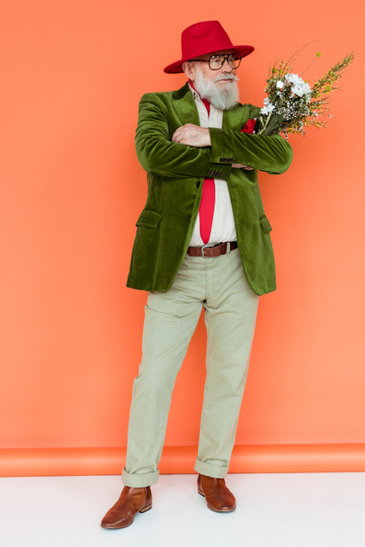 Full length of handsome elderly man with crossed arms holding wildflowers on white surface on coral background - Foto, afbeelding