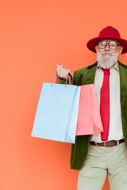 Handsome senior man in hat and eyeglasses holding shopping bags and looking at camera isolated on coral  - Foto, Imagem