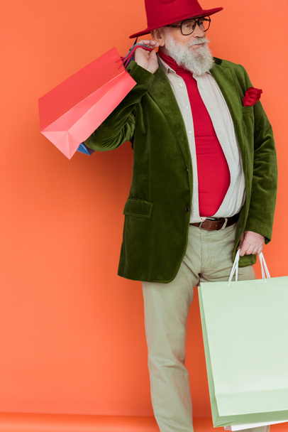 Fashionable elderly man holding shopping bags and looking away on coral background - Fotografie, Obrázek