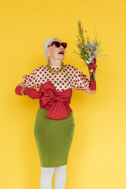 Fashionable elderly woman holding wildflowers isolated on yellow - Foto, immagini