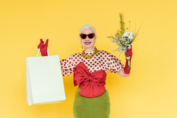 Mulher sênior elegante sorrindo enquanto segurando flores silvestres e sacos de compras no fundo amarelo
 - Foto, Imagem