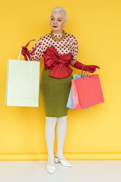 Full length of stylish senior woman with shopping bags and sunglasses looking away on white surface on yellow background - Fotografie, Obrázek