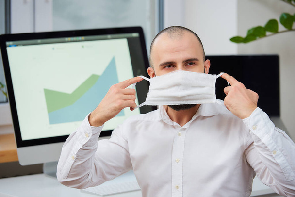 Mężczyzna zakładający medyczną maskę ochronną przeciwko koronawirusowi. Pracownik biurowy w swoim miejscu pracy z komputerami i zielonymi fabrykami w tle. kwarantanna 2019-nCoV. - Zdjęcie, obraz