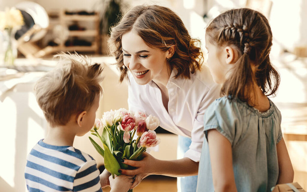 Feliz Día de las Madres! Niños niño y niña felicitar a la madre sonriente y darle flores ramo de tulipanes durante la celebración de vacaciones en la cocina en hom
 - Foto, Imagen