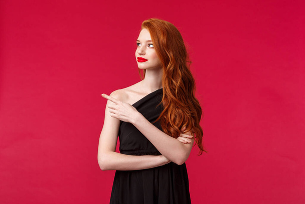 Celebrazione, eventi, concept moda. Ritratto di elegante giovane rossa sottile donna in abito nero elegante, indossare trucco rossetto rosso, guardare e puntare a sinistra con curioso volto interessato
 - Foto, immagini