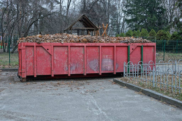 un gran contenedor de hierro rojo lleno de hojas secas se encuentra en el pavimento en un parque
 - Foto, imagen