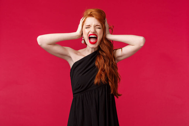 Portrait of distressed and pressured young redhead woman in black dress, screaming depressed losing control over emotions, hold hands on head in denial, found out bad truth, black background - Photo, Image